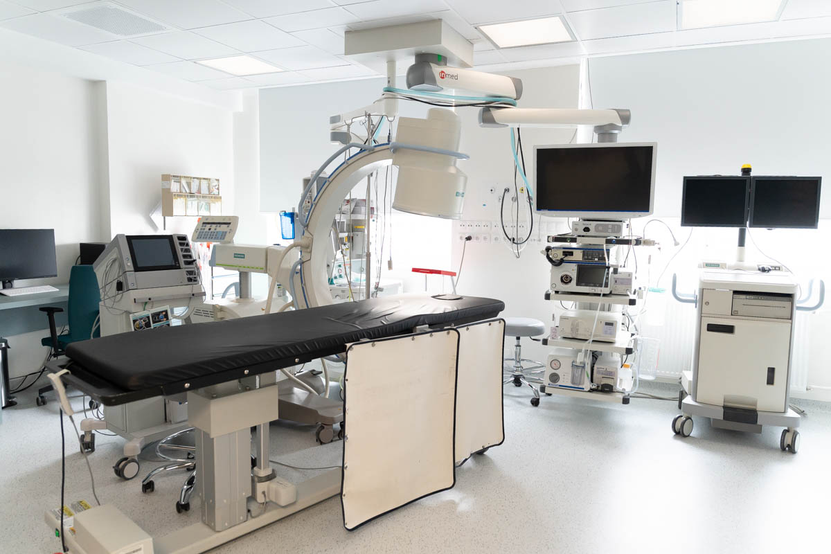 A modern medical examination room equipped with an operating table, imaging equipment, monitors, and various diagnostic and surgical devices.