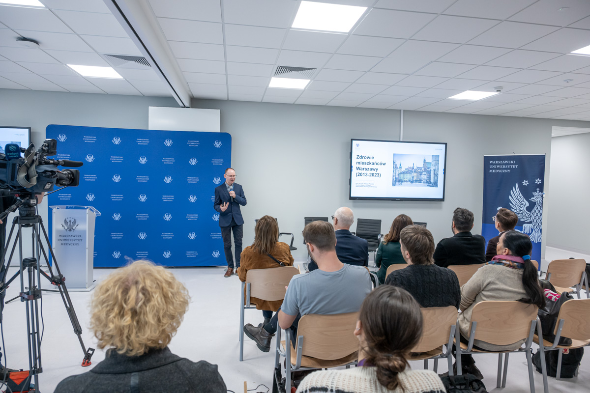 A speaker is standing in front of a group of about fifteen people and giving a presentation. He is showing a slide with a title related to the history of the inhabitants of Warsaw. The participants are seated in rows, facing the screen. On the left, there is a camera recording the event. In the background, boards with the institution’s logo are visible.
