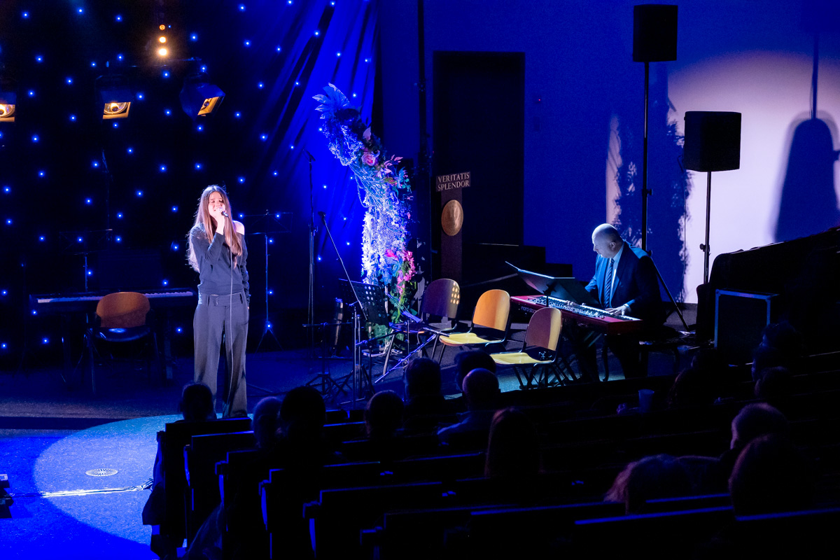 A woman is standing on stage and singing into a microphone. She is holding the microphone with both hands and looking straight ahead with a serious expression. On the right, a man is playing the keyboard, leaning over the instrument. The audience is seated in dim light in front of the stage.