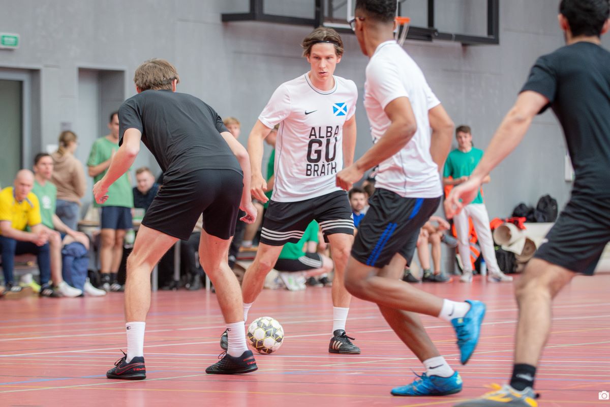 Players participating in an indoor football match are fighting for the ball in the center of the pitch, while the remaining participants and spectators watch the game from the sidelines.