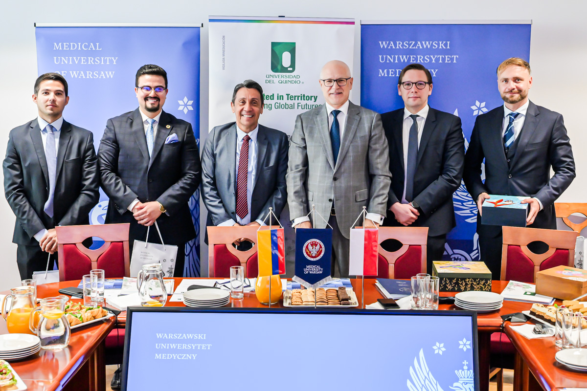 Six men are standing together, all dressed elegantly and smiling for the photo. In the background, three banners are hanging: blue banners with the WUM logo on the left and right, and a white banner from Columbia University in the center.