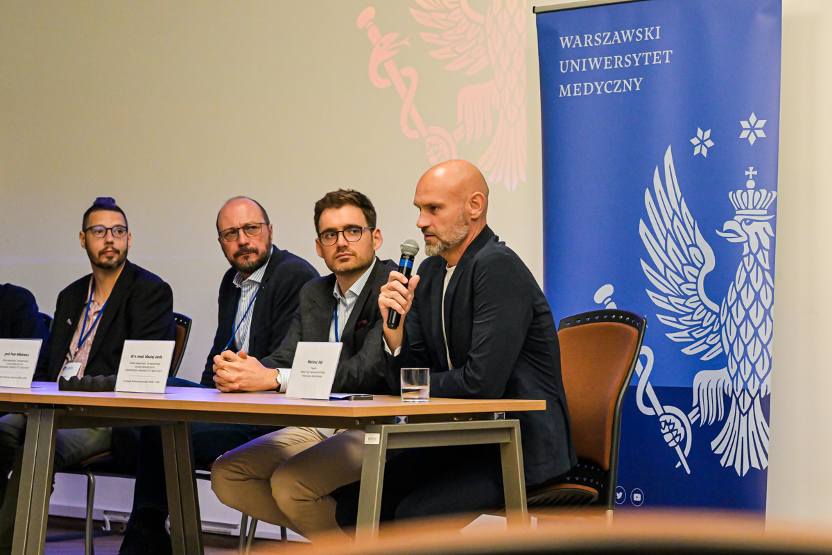 Four men dressed formally are sitting at a table. The man on the right is holding a microphone and speaking. The others are looking in his direction. Behind them is a blue banner with the WUM logo — a white eagle.