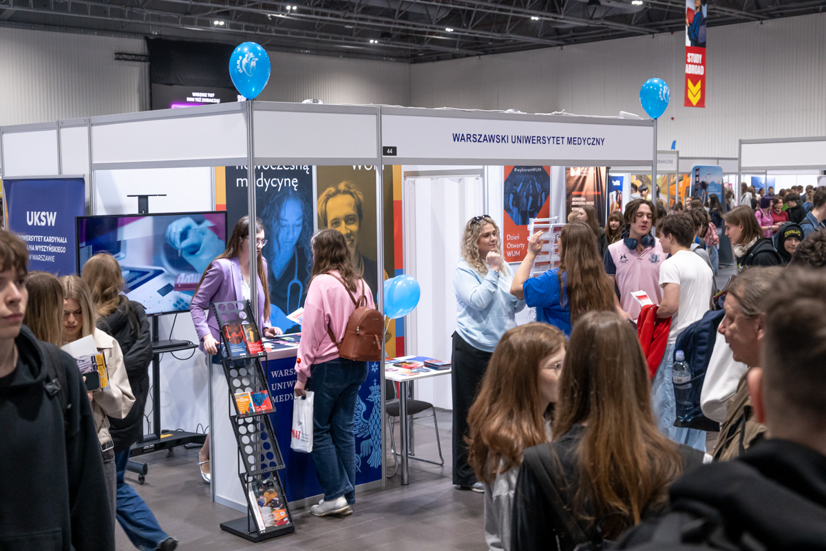 The trade hall features a booth of the Medical University of Warsaw, decorated in blue and displaying informational materials. Several people are looking at brochures and talking at the booth. In the background, other stands and visitors can be seen.