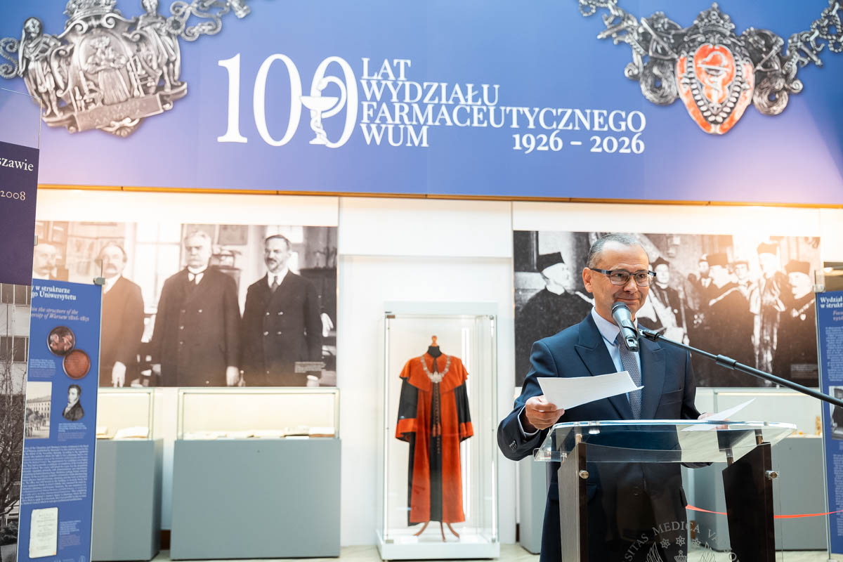 A speaker delivering an address during the ceremony marking the 100th anniversary of the WUM Faculty of Pharmacy, set against a backdrop of historical displays and showcases with university memorabilia.