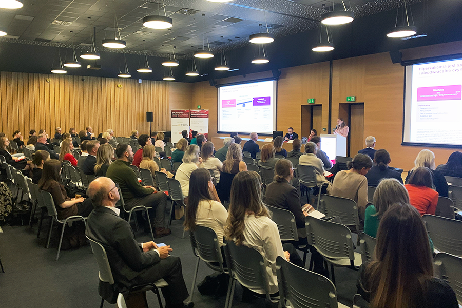 W dużej sali konferencyjnej siedzi kilkadziesiąt osób skierowanych w stronę sceny. Na scenie kilka osób siedzi przy stole, a jedna osoba stoi przy mównicy. Na dwóch dużych ekranach wyświetlane są slajdy w kolorach białych, różowych i niebieskich.