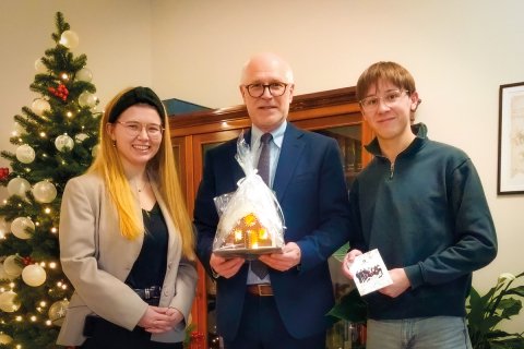 Three people — a woman and two men — are posing for a photo. The man in the middle is holding a gingerbread house. In the background, a decorated Christmas tree is visible.