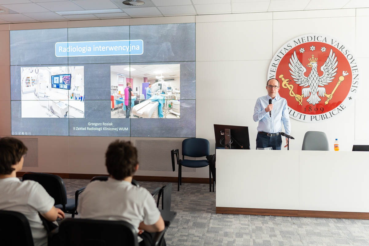 Prelegent stojący przy mównicy prowadzi prezentację na temat radiologii interwencyjnej, a na dużym ekranie za nim wyświetlane są zdjęcia sali zabiegowej. W pierwszym rzędzie siedzą słuchacze. Po prawej stronie widoczne jest godło uczelni medycznej.
