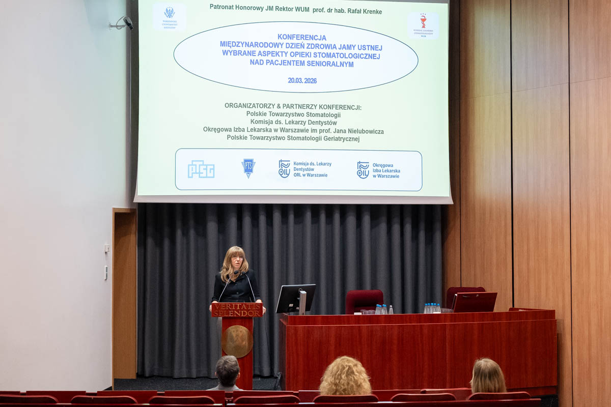 Osoba stojąca przy mównicy na tle dużego ekranu z prezentacją o immunosenescencji. Osoba ma jasne włosy, jasnoniebieską marynarkę i trzyma pilot do slajdów. Przed mównicą siedzą dwie osoby zwrócone w stronę sceny.