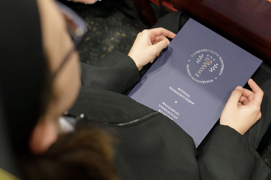 A person is sitting in an armchair and holding a closed navy-blue folder in their hands. The cover features the logo and the name of the Faculty of Pharmacy of the Medical University of Warsaw. The folder rests on their lap
