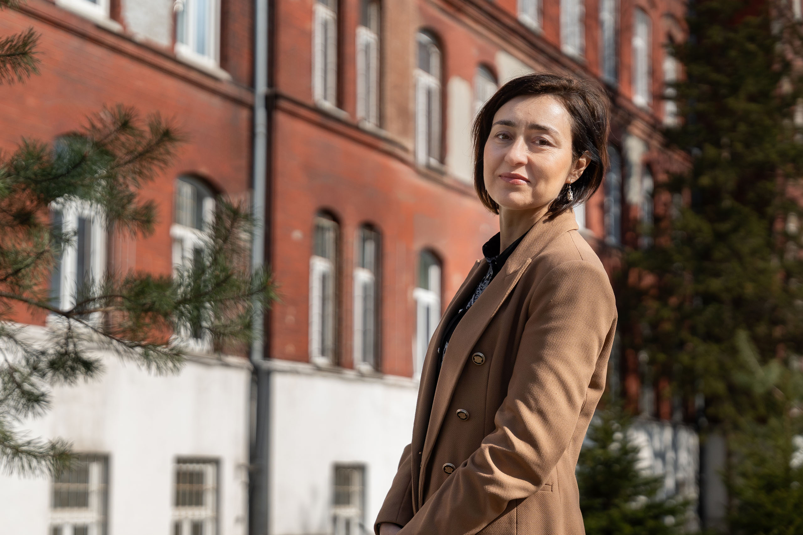 A person standing outdoors in front of a large red-brick building, wearing a beige coat. Trees and rows of windows are visible in the background.
