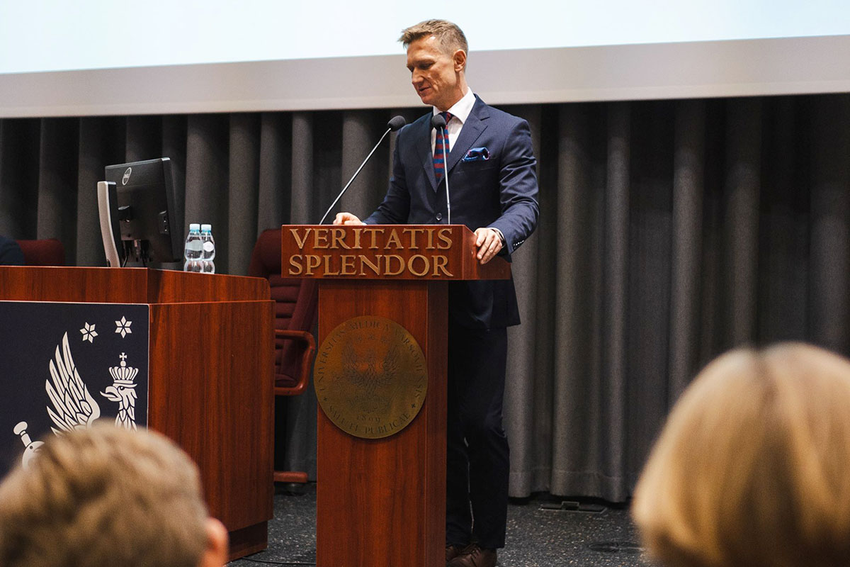 Osoba w garniturze przemawia z mównicy podczas wydarzenia akademickiego; na mównicy widoczne jest hasło Veritatis Splendor, a w tle znajduje się sala wykładowa i ekran projekcyjny.