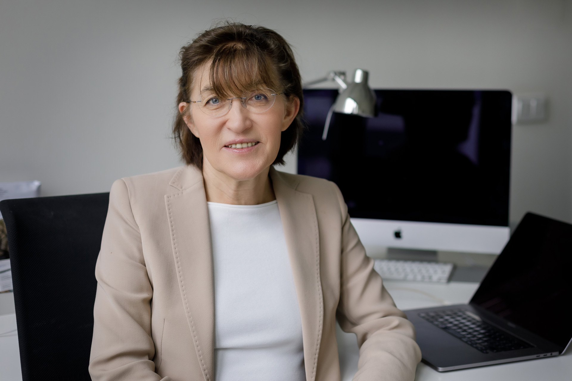 Profesor Hanna Szajewska siedzi na krześle przy biurku. Ma na sobie jasną marynarkę i białą bluzkę. W tle znajduje się komputer stacjonarny z dużym monitorem, lampka biurkowa na metalowym wysięgniku oraz otwarty laptop.