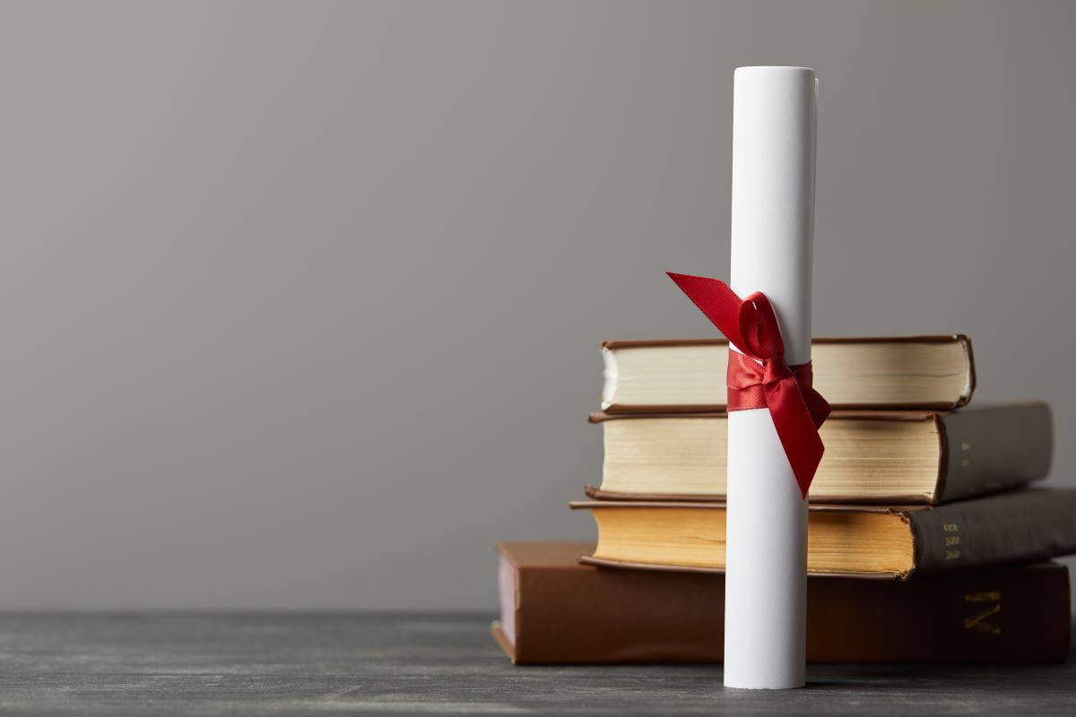 A rolled white parchment tied with a red ribbon, standing on a dark surface next to a stack of four hardcover books, set against a plain gray background.