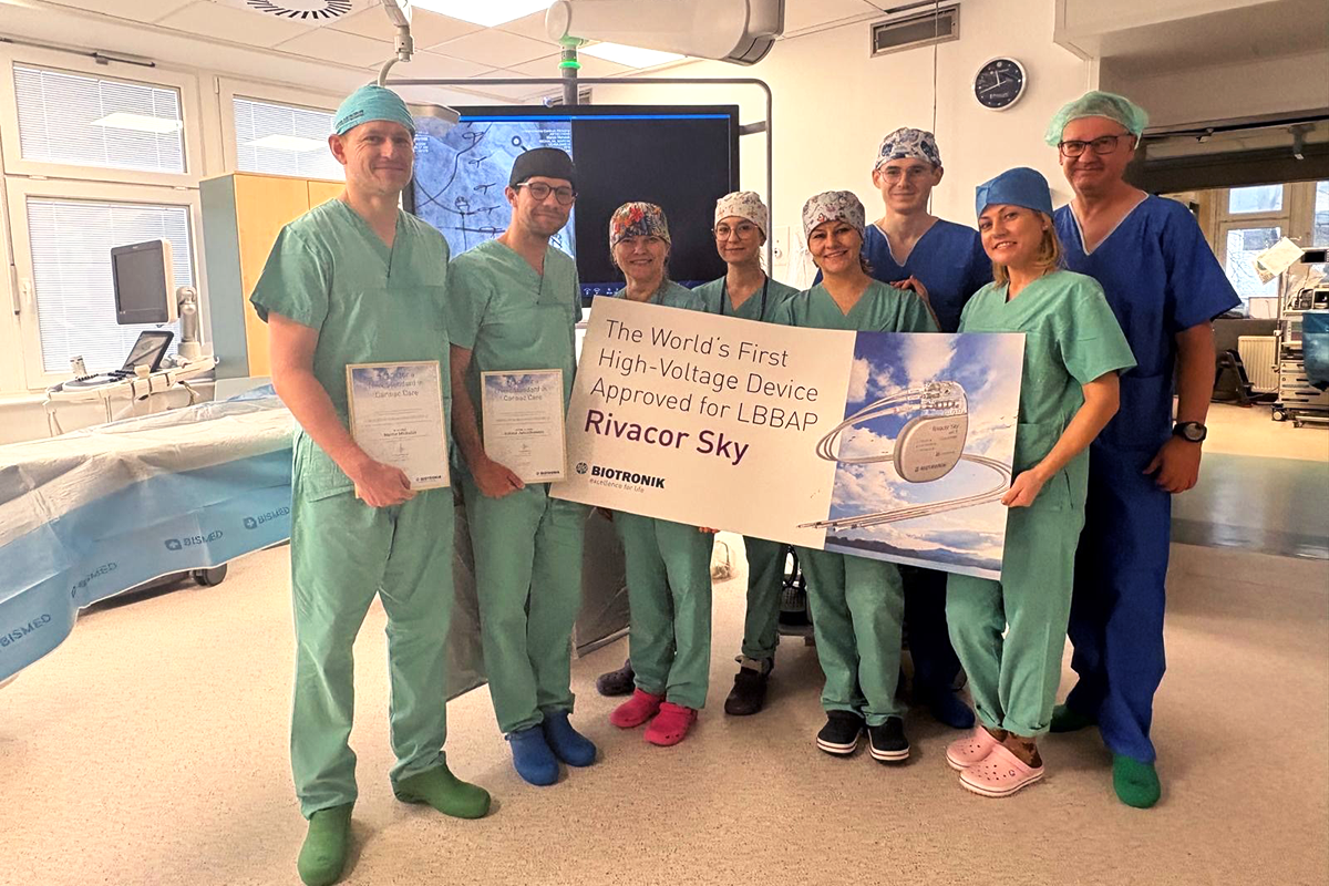 Zdjęcie przedstawia grupę ośmiu osób stojących w sali zabiegowej. Wszystkie osoby mają na sobie odzież medyczną: czepki, maseczki opuszczone pod brodę, fartuchy i ochraniacze na obuwie. Dwie osoby po lewej stronie trzymają dyplomy. Kilka osób po prawej stronie trzyma duży prostokątny baner. Na banerze znajduje się tekst: ‘The World's First High-Voltage Device Approved for LBBAP – Rivacor Sky’ oraz logo firmy Biotronik. W tle widać monitor z obrazem medycznym, lampy operacyjne i sprzęt zabiegowy.
