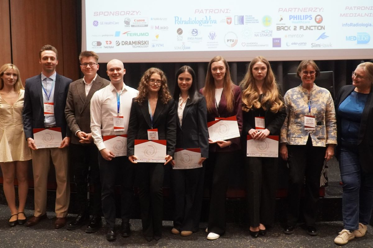 Grupa dorosłych uczestników stoi w rzędzie na scenie i trzyma certyfikaty. W tle widoczny jest ekran z nazwami sponsorów i partnerów wydarzenia, co wskazuje na uroczyste zakończenie konferencji lub szkolenia.