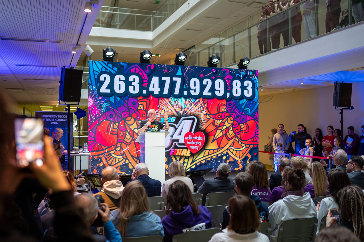 The interior of a large hall where a charity event is taking place. On the stage, a speaker stands at a lectern, and behind them there is a colorful graphic featuring the logo of the WOŚP Grand Finale and a large screen displaying the total amount raised: 263,477,929.83. In front of the stage, the audience is seated, while volunteers and observers are standing around.