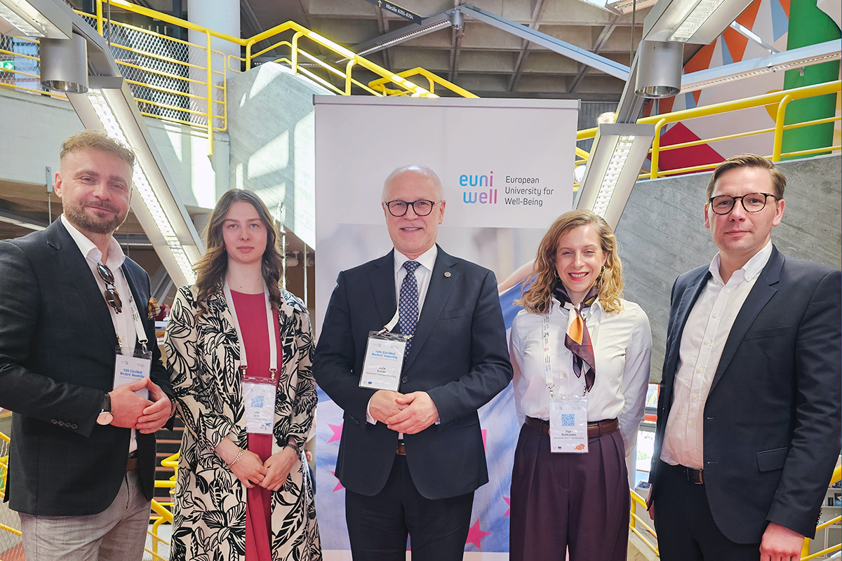 Five adults, three men and two women, stand side by side in an indoor space. They are dressed in formal clothes and wear conference badges on lanyards. Behind them there is a banner with the logo “European University for Well-Being” and yellow stair railings in a modern building interior.