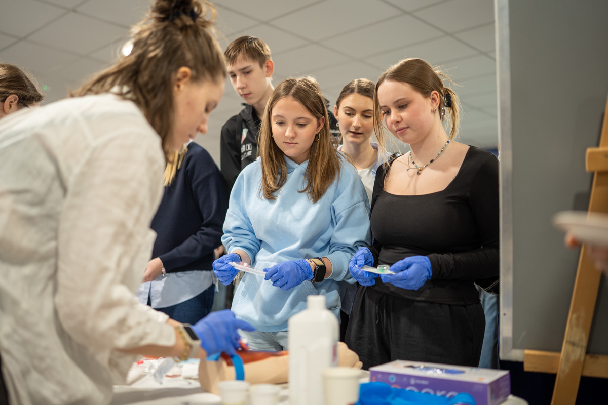 A group of young people stands around a workshop table while a facilitator demonstrates a procedure on a medical model; participants hold blue disposable gloves and small training tools in their hands.