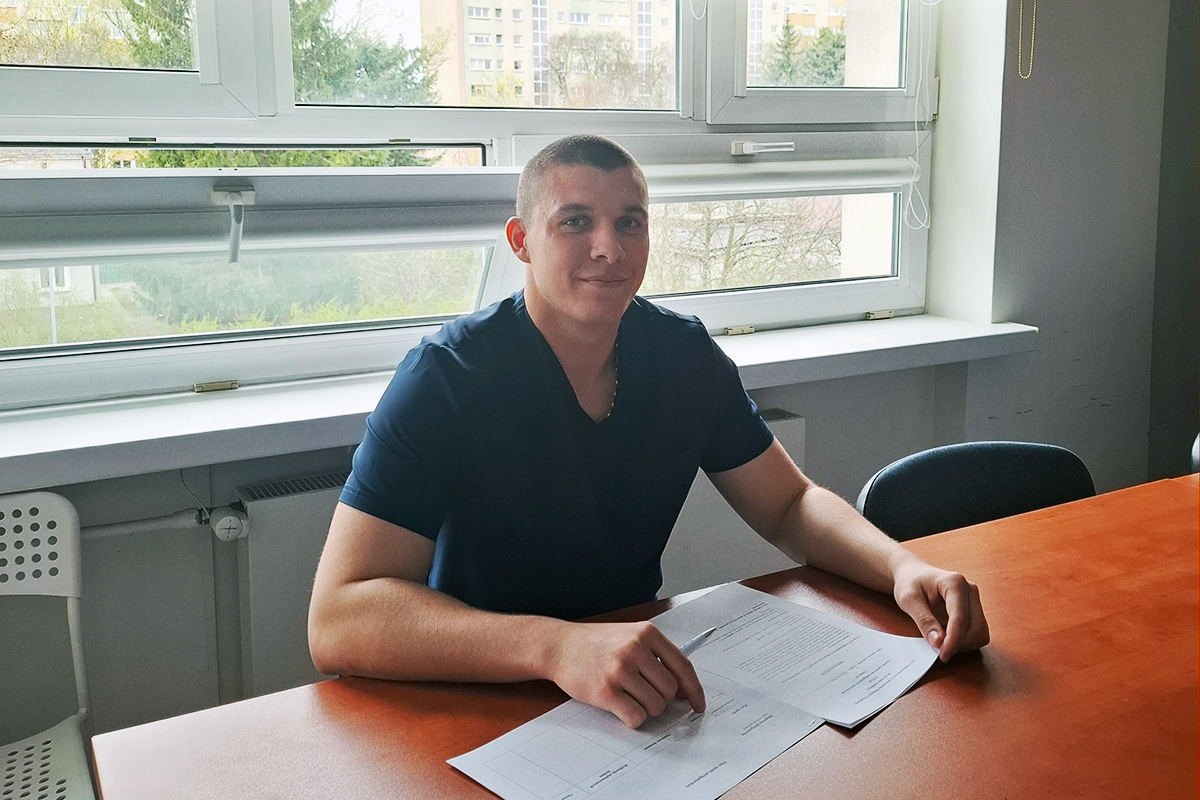 Młody chłopak siedzący za biurkiem, na którym widać kartki papieru. W tle, za nim, są okna.