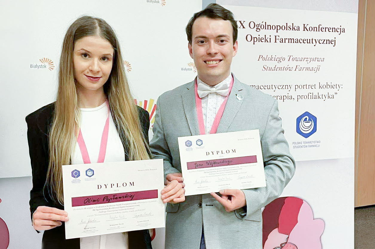 Dwie młode osoby - kobieta po lewej i mężczyzna po prawej - pozują do zdjęcia z dyplomami w rękach. Uśmiechają się. Za nimi stoi ścianka z informacjami o wydarzeniu.