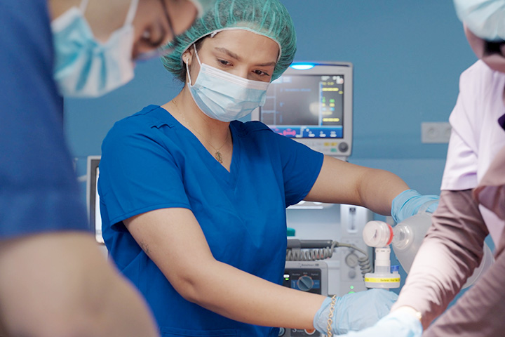 Grupa młodych osób w czasie zajęć symulacyjnych. Ubrani są w niebieskie fartuchy, na twarzach mają maseczki, na głowach czepki. W tle widać aparaturę medyczną.