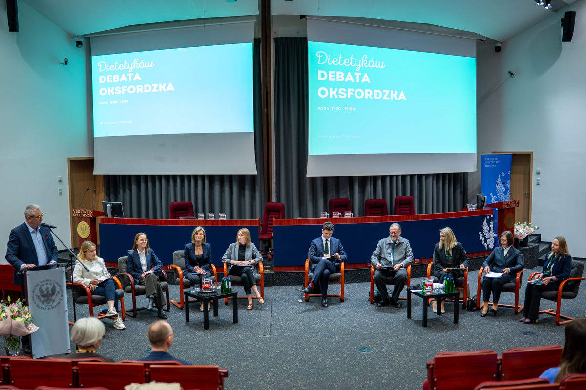Na sali konferencyjnej odbywa się panelowa debata oksfordzka. Kilkanaście osób siedzi w półokręgu na scenie, przed nimi stoliki z wodą. W tle widoczny jest ekran z napisem „Dietetyka. Debata oksfordzka”, a na pierwszym planie fragment widowni.