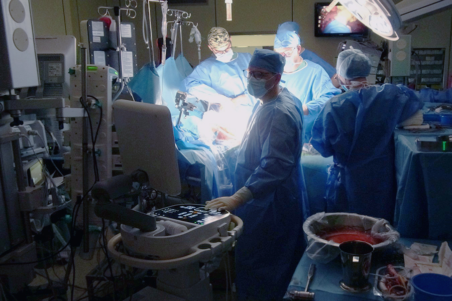 Sala operacyjna podczas trwającego zabiegu chirurgicznego. Kilka osób w sterylnych niebieskich fartuchach pracuje przy pacjencie, oświetlonym lampą operacyjną. Wokół znajdują się aparatura medyczna, monitory oraz stoliki z narzędziami.