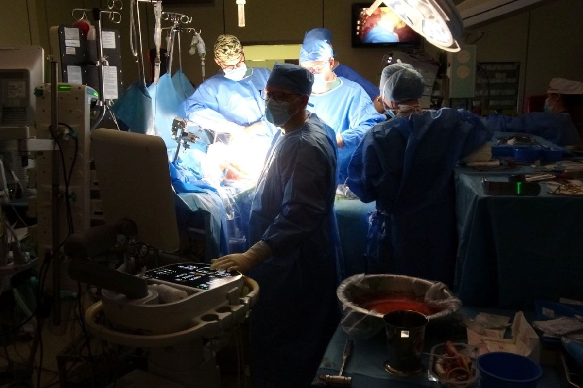Sala operacyjna podczas trwającego zabiegu chirurgicznego. Kilka osób w sterylnych niebieskich fartuchach pracuje przy pacjencie, oświetlonym lampą operacyjną. Wokół znajdują się aparatura medyczna, monitory oraz stoliki z narzędziami.
