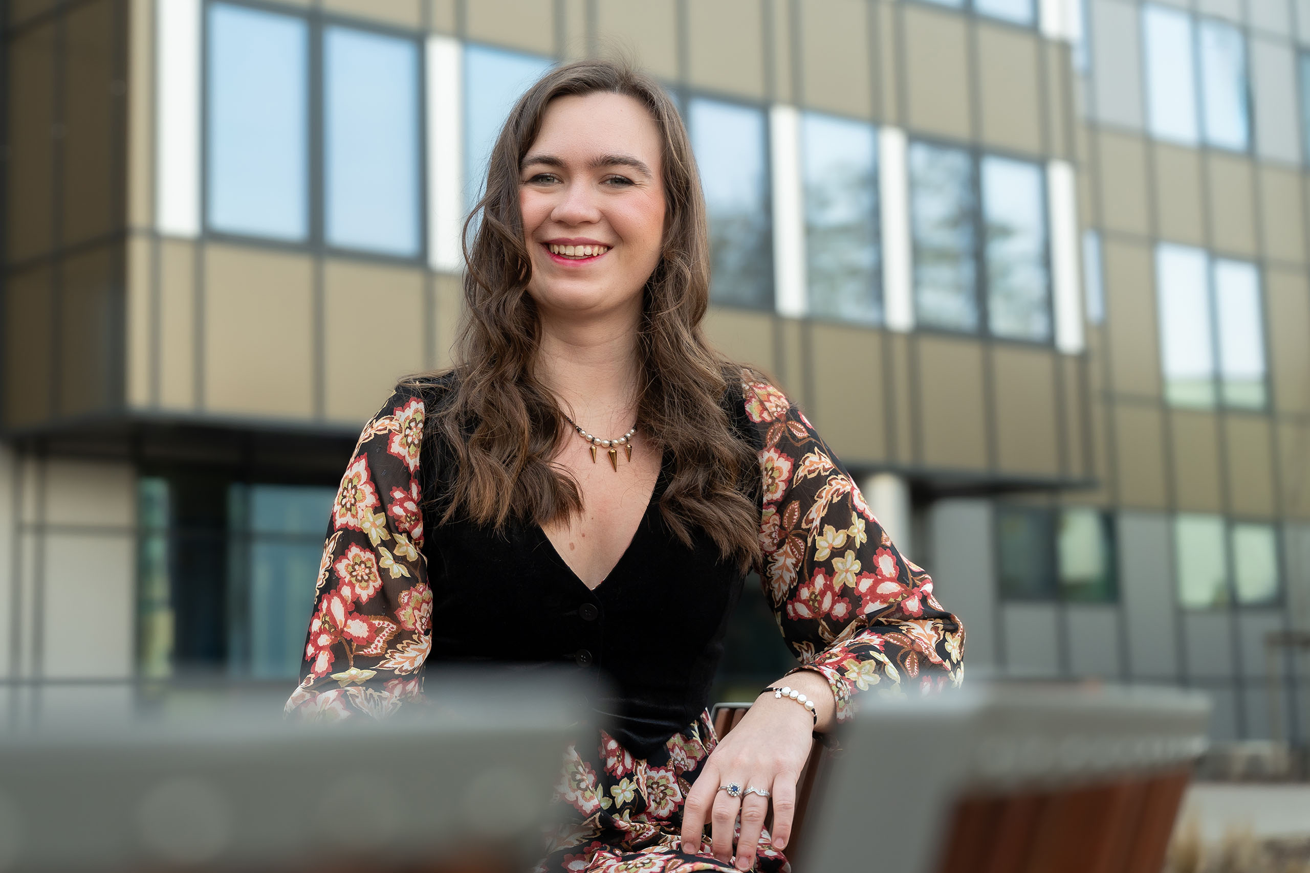 Młoda kobieta uśmiecha się do zdjęcia. Siedzi na ławce. W tle widać budynek z wysokimi oknami.