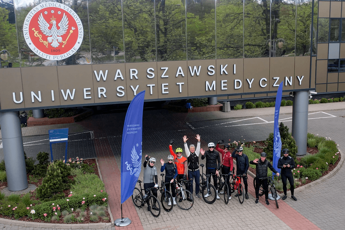 Grupa osób pozuje do zdjęcia. Większość jest ubrana w stroje kolarskie, na głowie ma kaski, są z rowerami. Stoją przy nowoczesnym budynku z oszkloną fasadą. Na budynku widać godło WUM oraz nazwę: Warszawski Uniwersytet Medyczny.