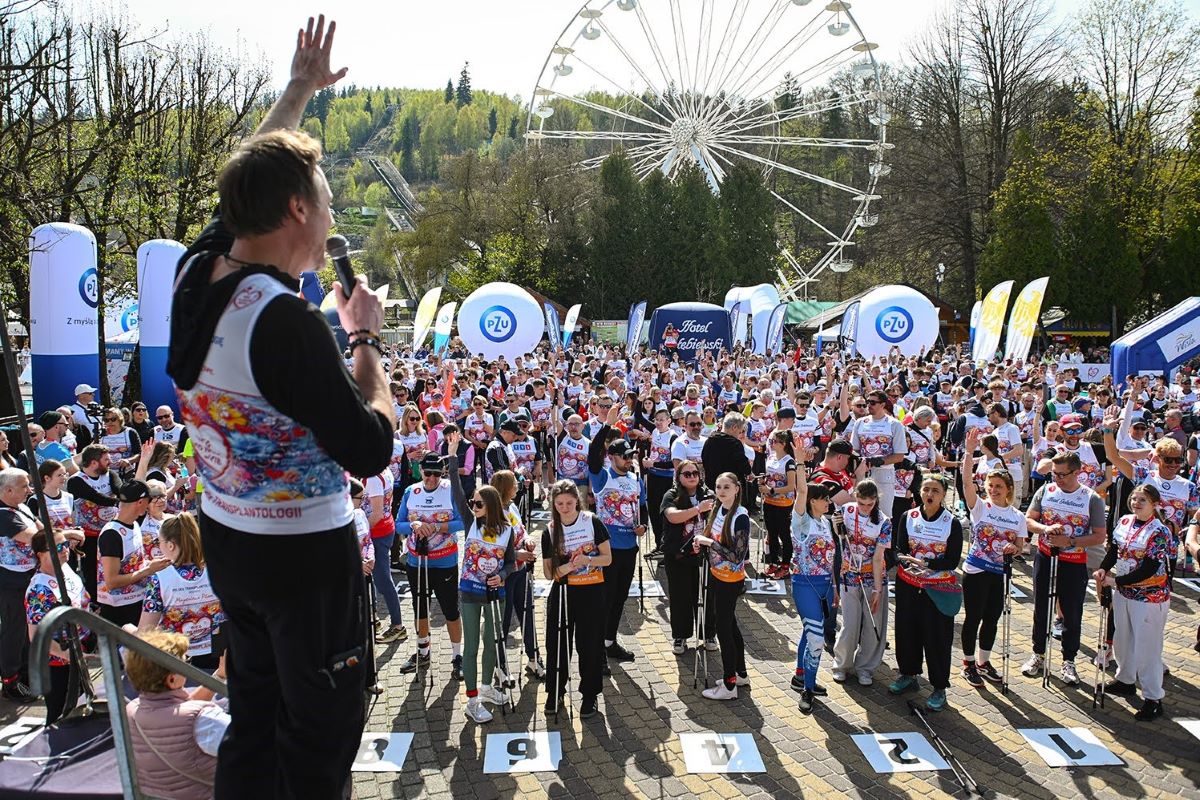 Osoba stojąca na podwyższeniu trzyma mikrofon w jednej ręce i unosi drugą rękę, zwracając się do licznego tłumu uczestników wydarzenia sportowego na otwartej przestrzeni. Uczestnicy stoją ciasno przed sceną, wielu trzyma w dłoniach kijki do nordic walking i ma na sobie jednakowe koszulki z kolorowymi nadrukami oraz numerami startowymi na piersi. Na placu przed sceną widoczne są oznaczenia pól startowych z numerami. W tle znajdują się banery i balony reklamowe, namioty eventowe, drzewa.