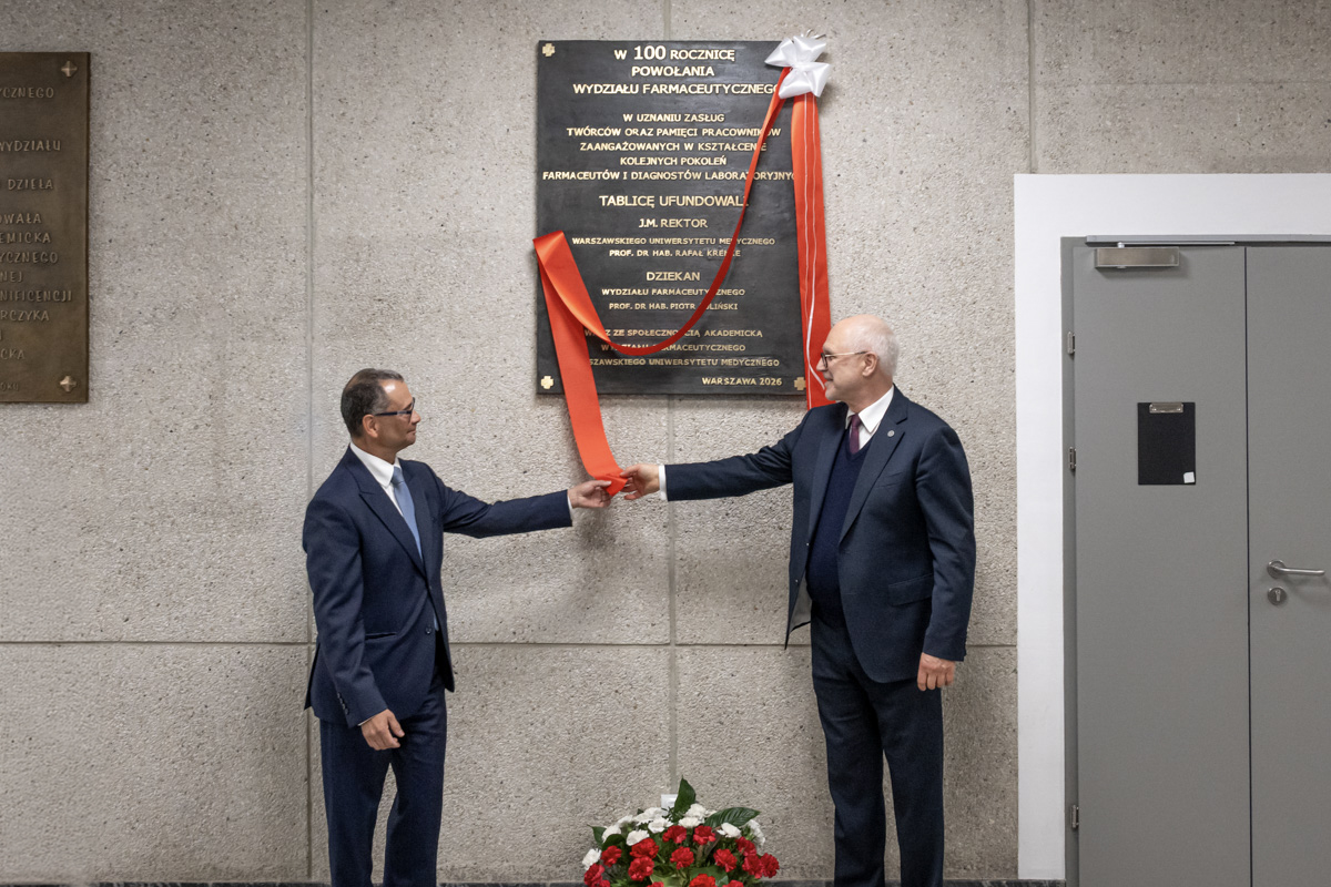 Dwie osoby w formalnych garniturach stoją przy ścianie z pamiątkową tablicą. Każda z osób trzyma w dłoni koniec czerwonej wstęgi, którą odsłaniają tablicę. Pod tablicą ustawiony jest kosz z biało‑czerwonymi kwiatami.