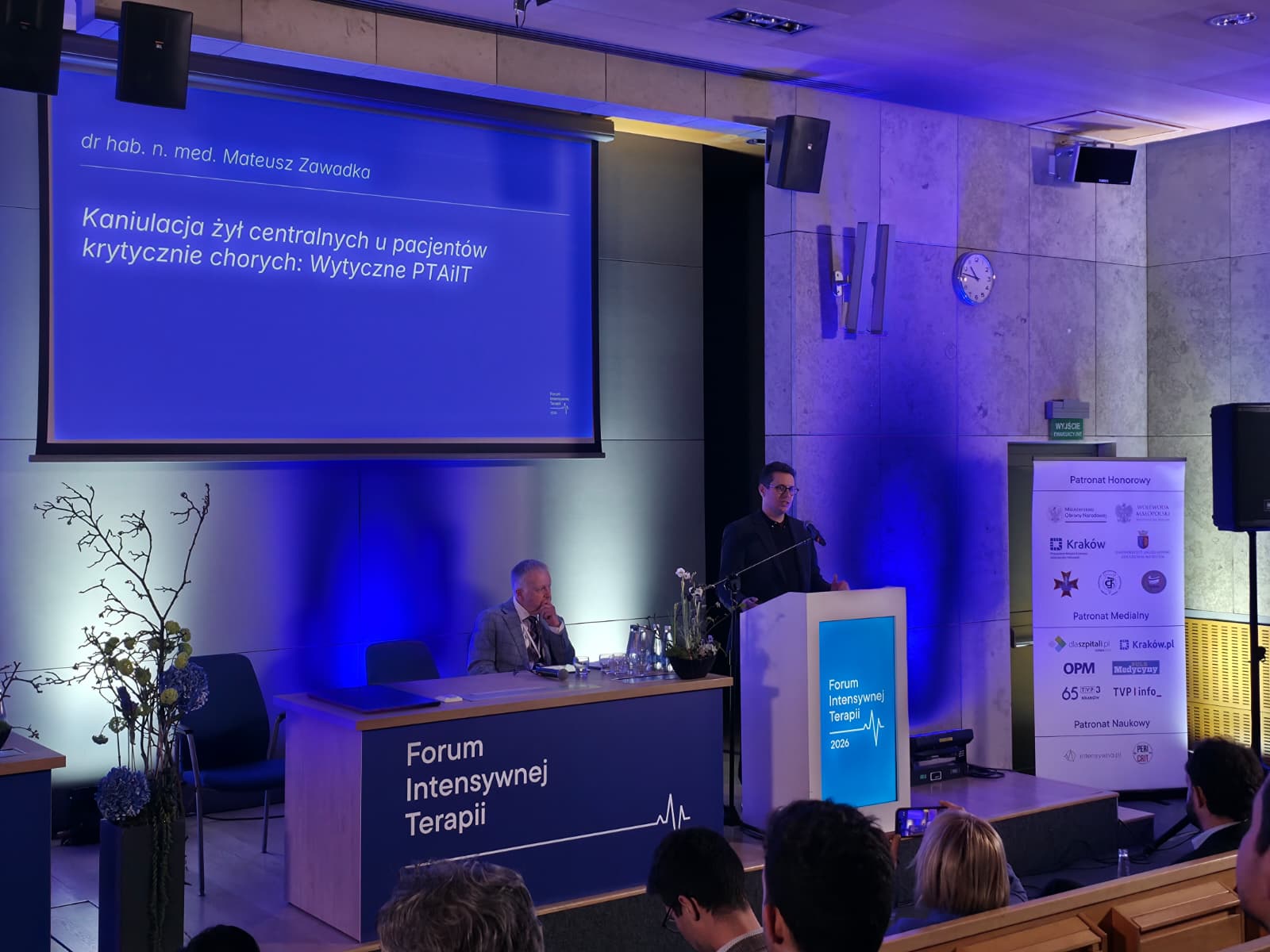 Sala konferencyjna podczas wydarzenia naukowego. Na scenie widoczny jest prelegent stojący przy mównicy oraz druga osoba siedząca przy stole prezydialnym. Za nimi wyświetlana jest prezentacja z tytułem dotyczącym kaniulacji żył centralnych u pacjentów krytycznie chorych. Na pierwszym planie znajduje się publiczność, a scena jest oświetlona niebieskim światłem.