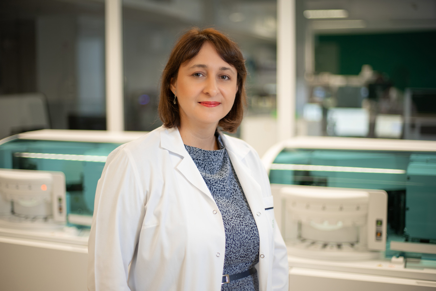 Jedna osoba stoi w laboratorium diagnostycznym i patrzy w stronę aparatu. Ma na sobie biały fartuch laboratoryjny. Za nią znajdują się nowoczesne analizatory medyczne ustawione w rzędzie. Urządzenia stoją za przeszkloną osłoną, a przestrzeń ma jasne, sterylne oświetlenie.