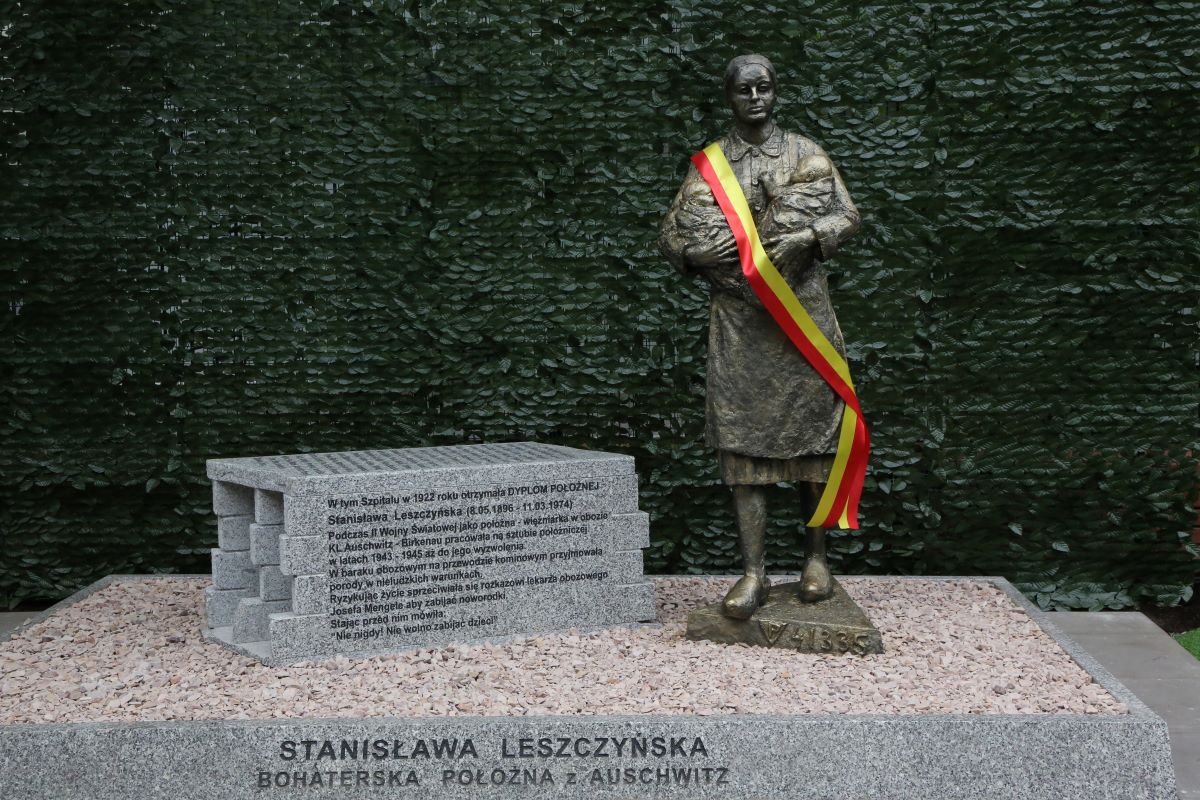 Pomnik Stanisławy Leszczyńskiej przedstawiający postać trzymającą niemowlę owinięte w materiał. Na pomniku zawieszona jest biało‑czerwona wstęga. Przed pomnikiem znajduje się kamienny postument z inskrypcją, a tło stanowi zielona ściana roślin.