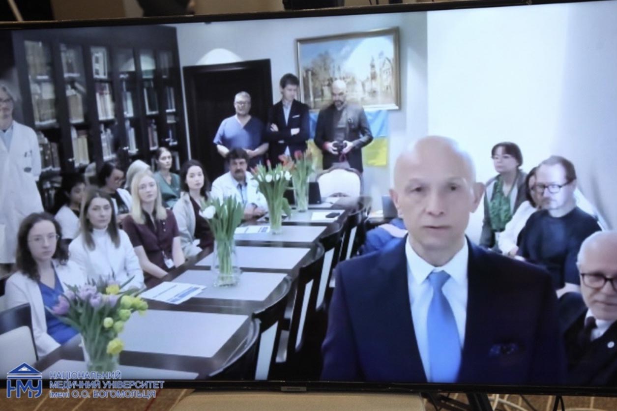 A conference room with a long table and several people seated on both sides. At the front, one person in a suit stands facing the camera. On the table there are vases with tulip flowers and printed documents. Bookshelves and a framed picture are in the background.