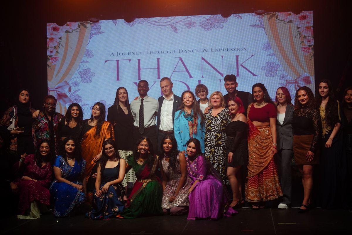 A large group of performers standing on a stage in colorful traditional and formal costumes, posing in front of a screen with the word “THANK YOU.”