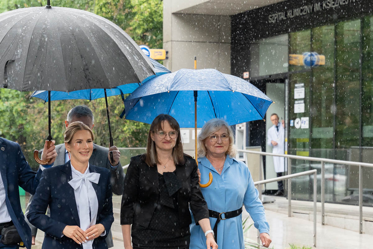 Several adults walk in the rain outside a hospital entrance, holding umbrellas. The hospital building and signage are visible in the background.