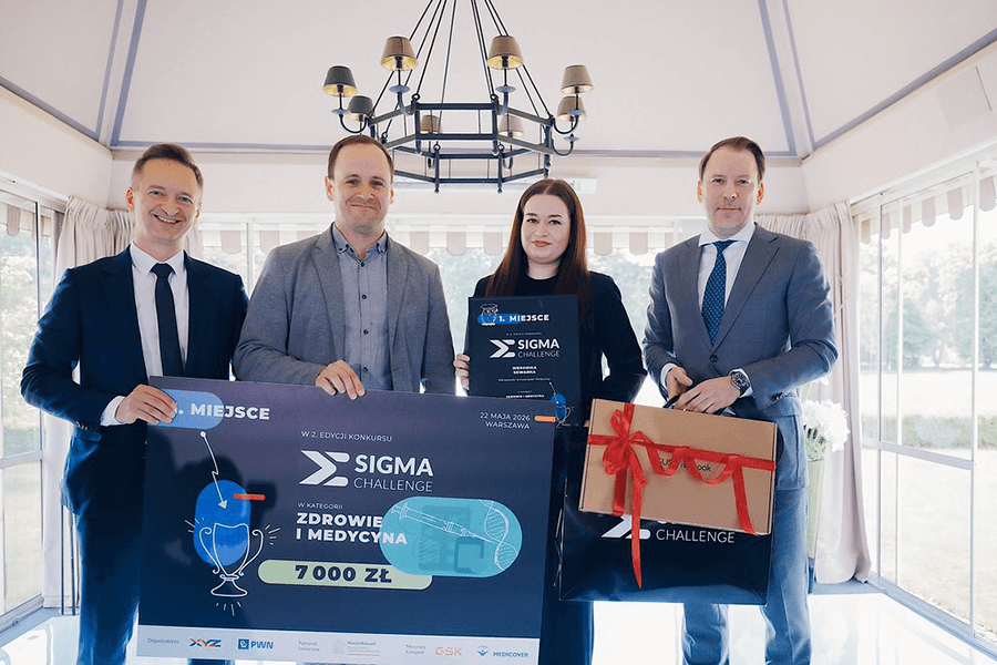 Three men and a woman are posing for a group photo. Two of the men are holding a large check, the woman is holding a framed diploma, and the third man is holding a gift package.