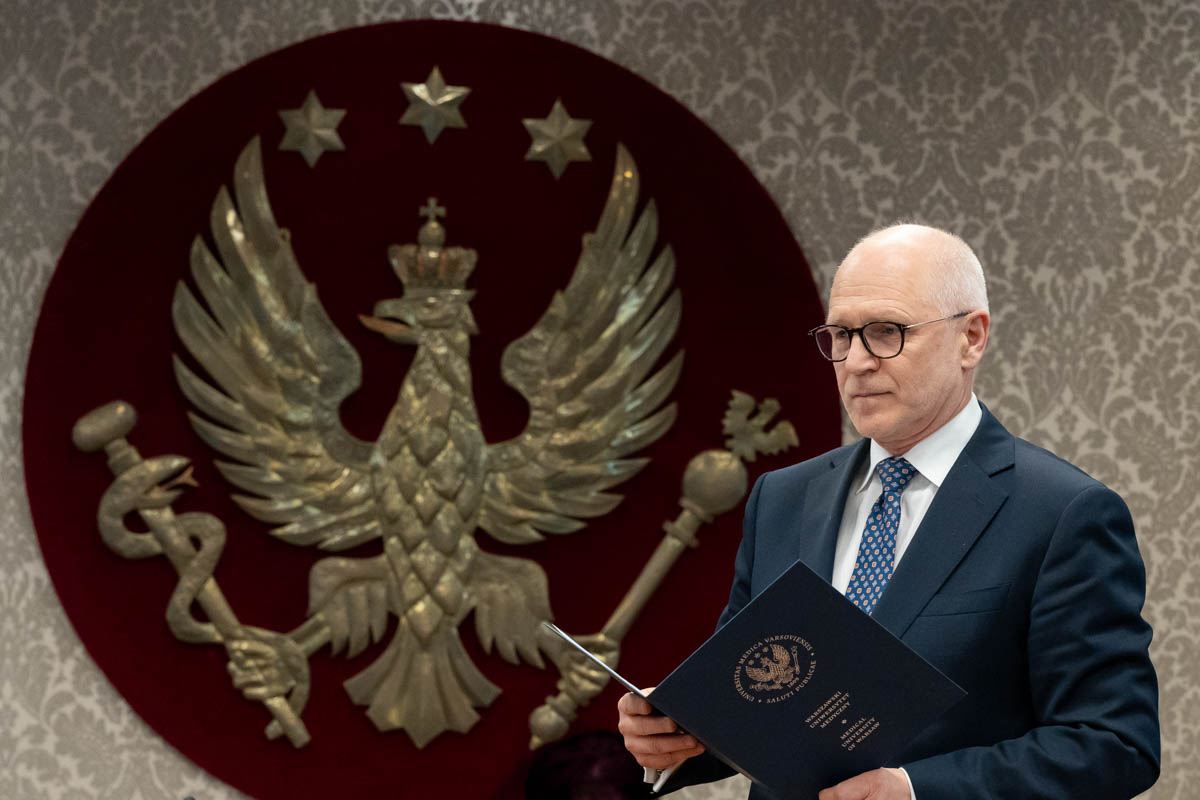 Mężczyzna stoi przy mównicy i odczytuje dokument. W tle widoczny jest herb uczelni - złoty orzeł na czerwonym tle. 