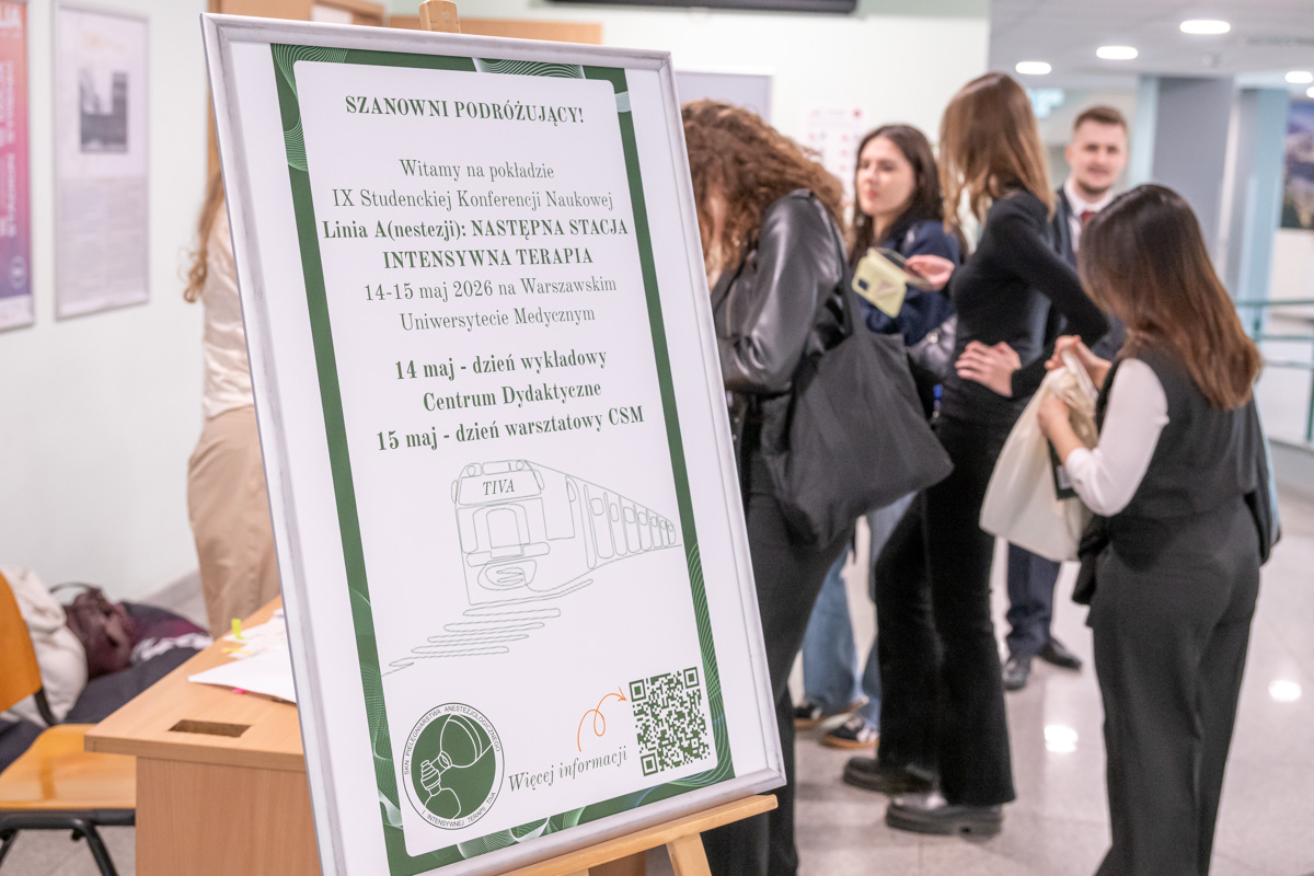Tablica informacyjna na stojaku z napisem dotyczącym IX Studenckiej Konferencji Naukowej „Linia A (anestezja): Następna stacja intensywna terapia”, ustawiona w holu. W tle kilka osób stoi i rozmawia, część trzyma torby oraz materiały konferencyjne.