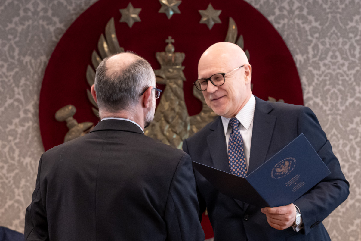 Dwóch mężczyzn stoi naprzeciw siebie w sali senatu WUM. Jeden wręcza drugiemu otwartą teczkę z dokumentem. W tle widoczny jest herb uczelni.