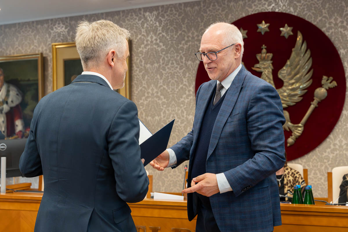 Dwóch mężczyzn stoi naprzeciw siebie w sali senatu WUM. Jeden wręcza drugiemu otwartą teczkę z dokumentem. W tle widoczny jest herb uczelni.