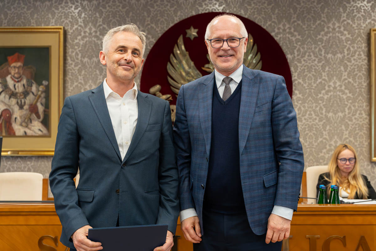 Dwóch mężczyzn stoi obok siebie w sali senatu WUM. Mężczyzna po lewej trzyma zamkniętą teczkę z dokumentem. Obaj mężczyźni uśmiechają się do zdjęcia. W tle siedzą uczestnicy posiedzenia oraz widoczny jest herb uczelni.
