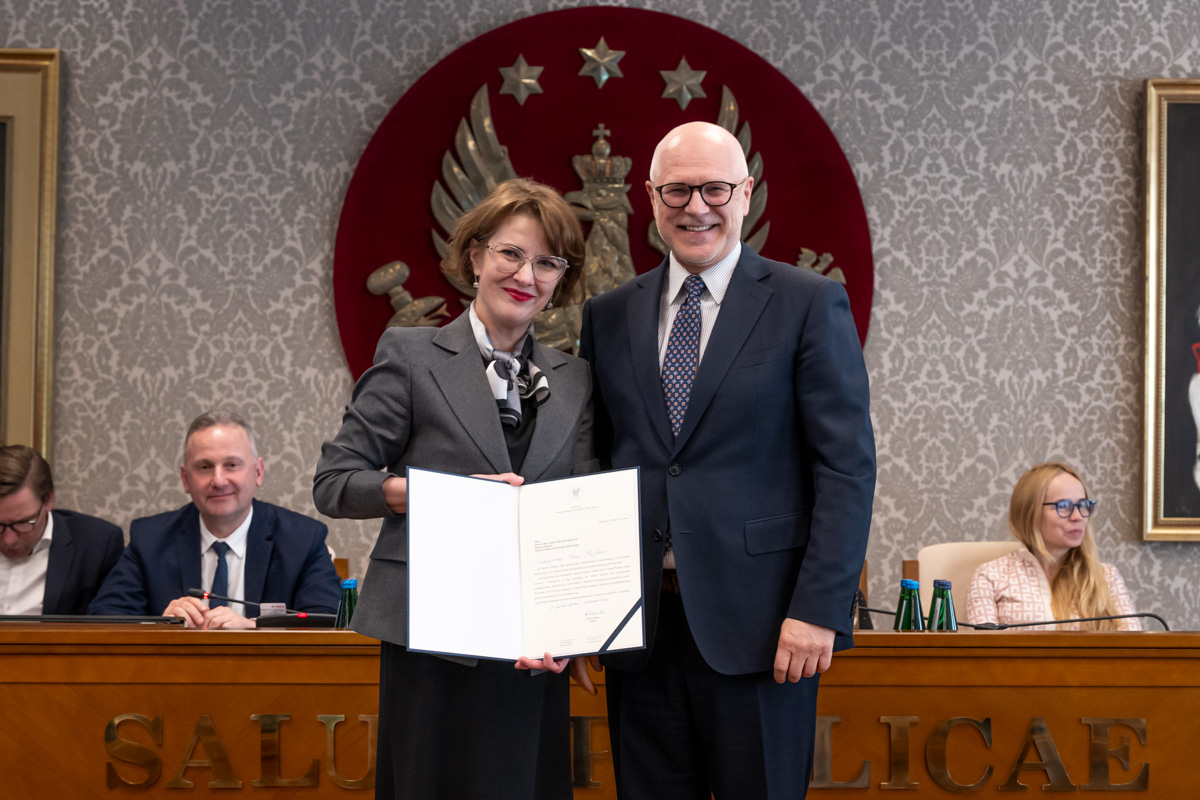 Kobieta i mężczyzna stoją obok siebie w sali senatu WUM. Kobieta trzyma otwartą teczkę z dokumentem. Obie osoby uśmiechają się do zdjęcia. W tle siedzą uczestnicy posiedzenia oraz widoczny jest herb uczelni.