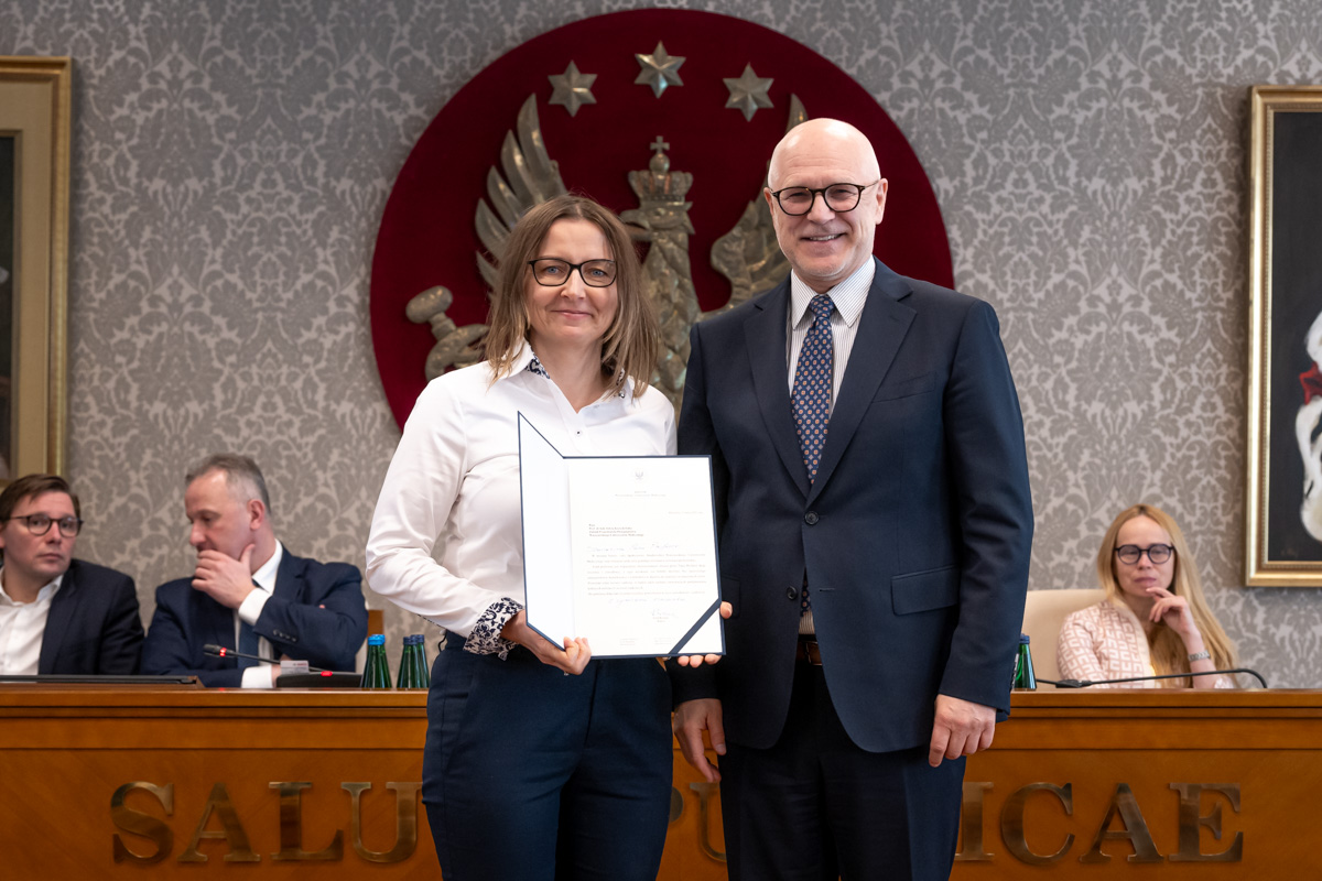 Kobieta i mężczyzna stoją obok siebie w sali senatu WUM. Kobieta trzyma otwartą teczkę z dokumentem. Obie osoby uśmiechają się do zdjęcia. W tle siedzą uczestnicy posiedzenia oraz widoczny jest herb uczelni.