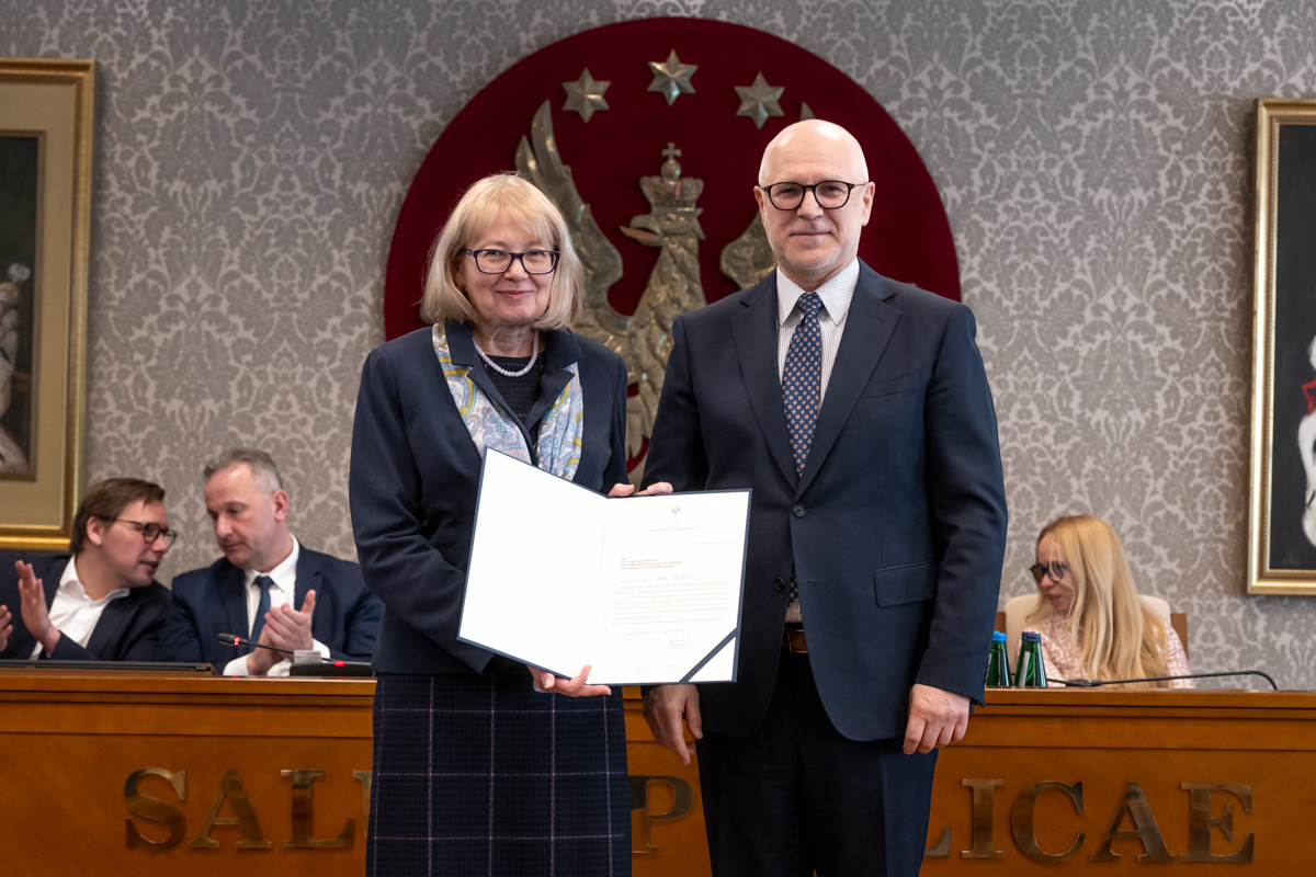 Kobieta i mężczyzna stoją obok siebie w sali senatu WUM. Kobieta trzyma otwartą teczkę z dokumentem. Obie osoby uśmiechają się do zdjęcia. W tle siedzą uczestnicy posiedzenia oraz widoczny jest herb uczelni.