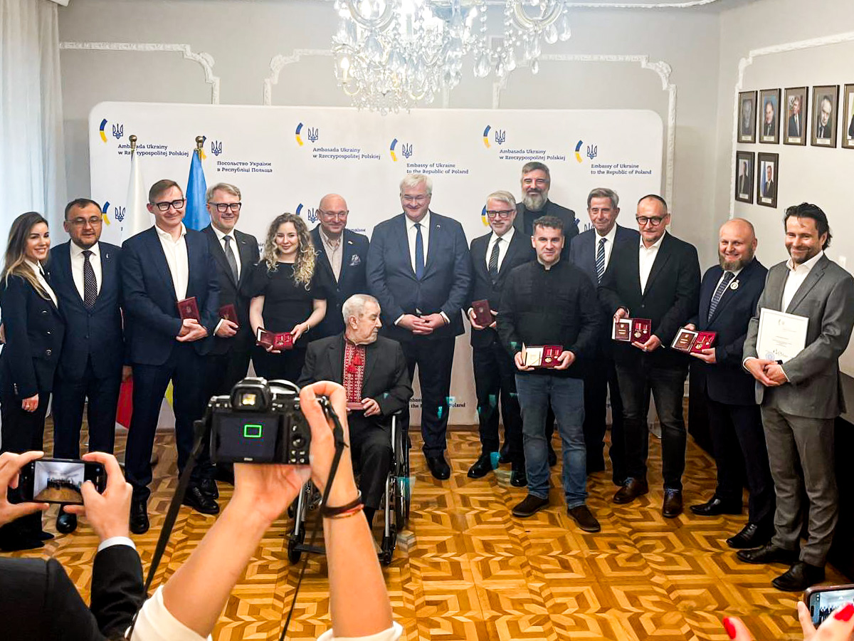 Grupa kilkunastu osób stojących w eleganckim pomieszczeniu podczas uroczystości. W centrum znajduje się osoba siedząca na wózku inwalidzkim, trzymająca dokument. Pozostałe osoby stoją w rzędzie i trzymają w rękach czerwone etui z odznaczeniami lub dyplomy. W tle znajduje się ścianka z napisem „Embassy of Ukraine in the Republic of Poland” oraz flagi. Na pierwszym planie widoczne są ręce osoby fotografującej i aparat fotograficzny.