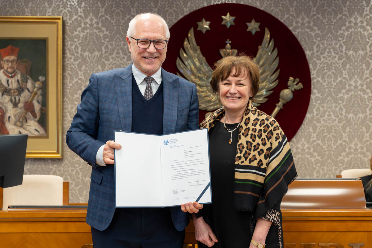 Kobieta i mężczyzna stoją obok siebie w sali senatu WUM. Kobieta trzyma otwartą teczkę z dokumentem. Obie osoby uśmiechają się do zdjęcia. W tle siedzą uczestnicy posiedzenia oraz widoczny jest herb uczelni.
