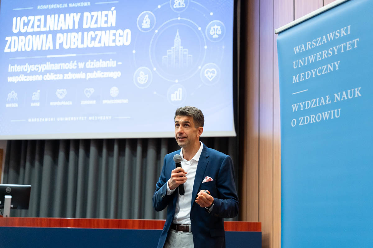 Mężczyzna z mikrofonem w czasie prezentacji. W tle widać ekran multimedialny ze slajdem informującym o konferencji, obok widać niebieski baner z nazwą Wydziału Nauk o Zdrowiu.